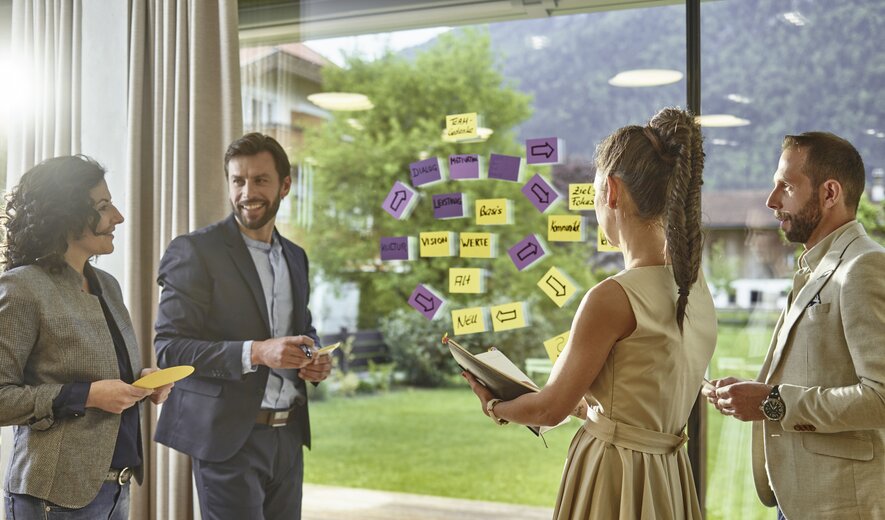 Eine vierköpfige Gruppe hält einen kreativen Workshop vor einer Glaswand voller bunter Haftnotizen.