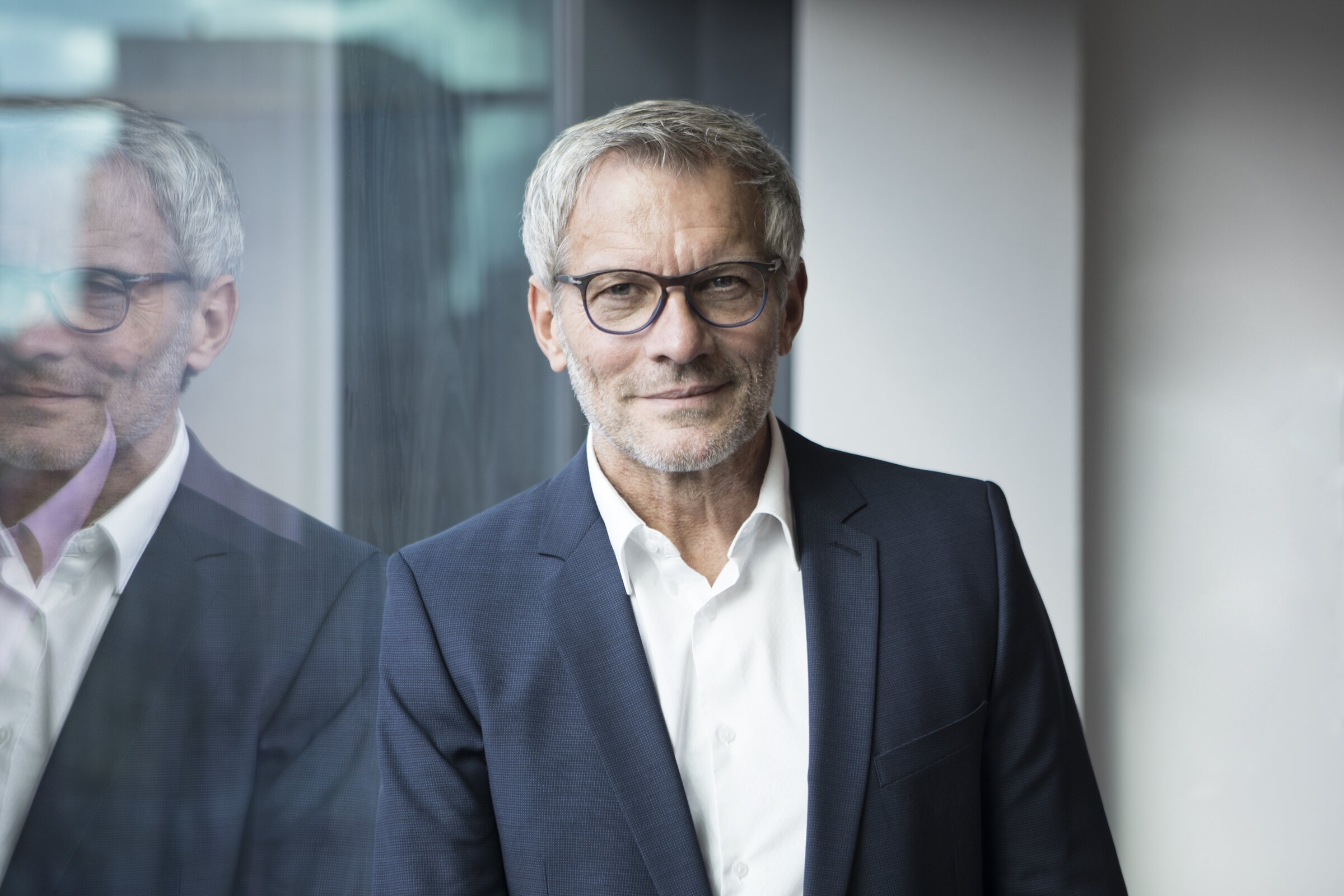 Ein Mann im Business-Anzug steht ruhig und selbstbewusst vor einer Glaswand.