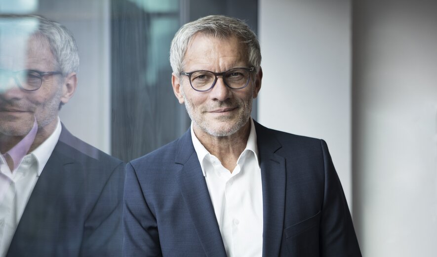 Ein Mann im Business-Anzug steht ruhig und selbstbewusst vor einer Glaswand.