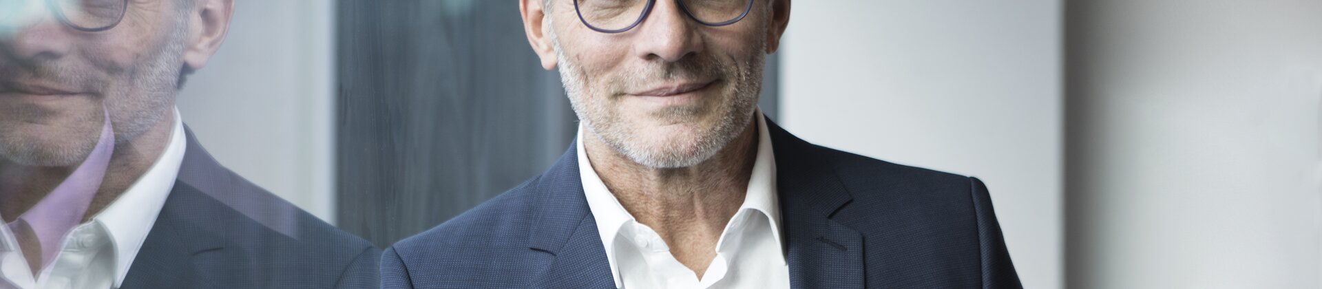 Ein Mann im Business-Anzug steht ruhig und selbstbewusst vor einer Glaswand.