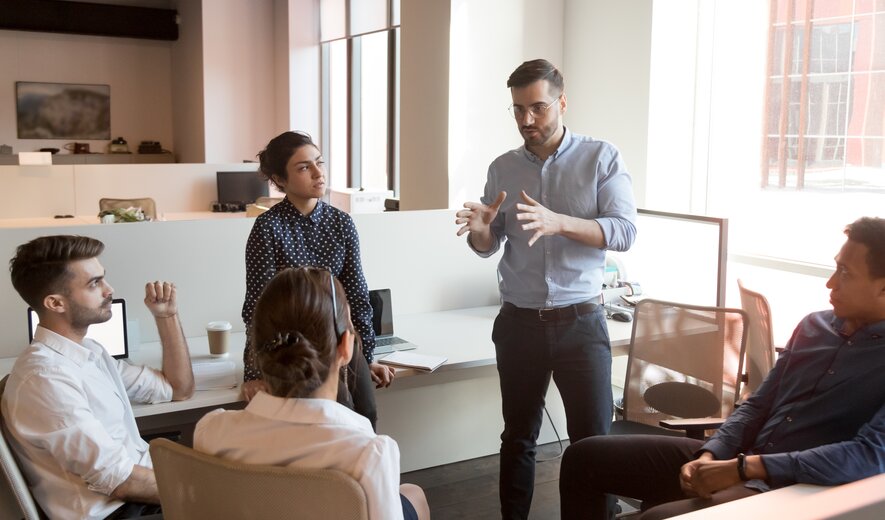 Ein Team sitzt in lockerer Runde zusammen, während ein stehender Kollege engagiert eine Idee erklärt.