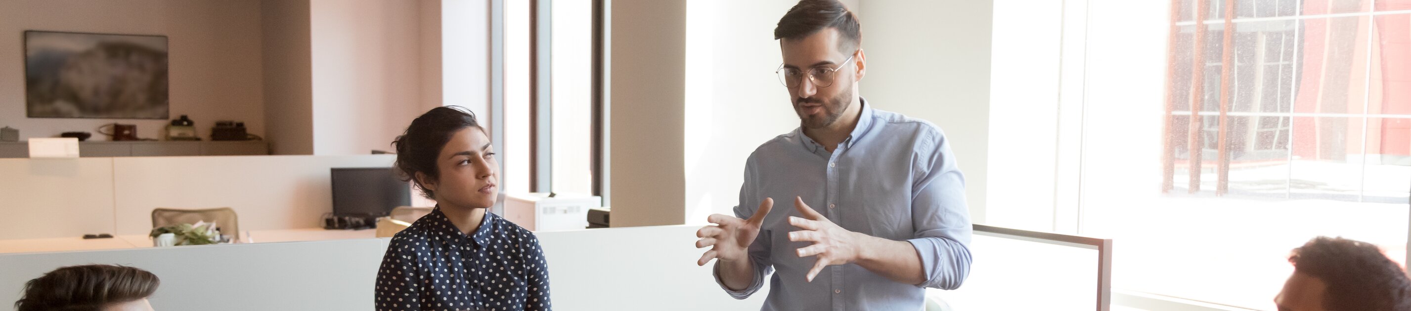 Ein Team sitzt in lockerer Runde zusammen, während ein stehender Kollege engagiert eine Idee erklärt.