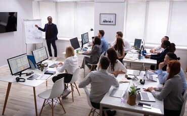 Eine Gruppe von Menschen sitzt gemeinsam an einem Tisch und arbeitet konzentriert an Laptops und Unterlagen – Symbol für Training, Wissensvermittlung und Zusammenarbeit im Seminar.