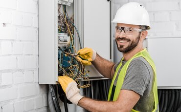 Ein Elektriker arbeitet lächelnd an einem offenen Schaltschrank voller Kabel.
