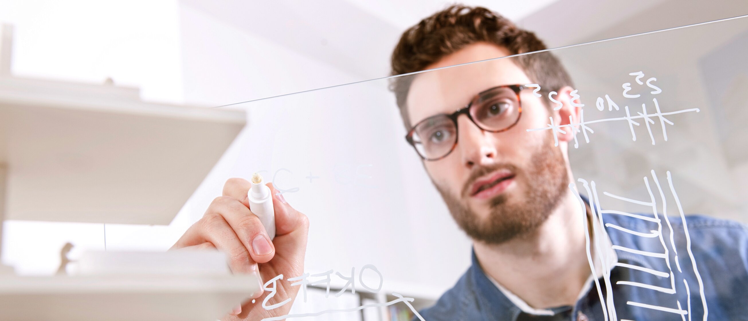 Mann schreibt technische Formeln auf Glaswand – Symbol für technische Weiterbildung und Innovation.