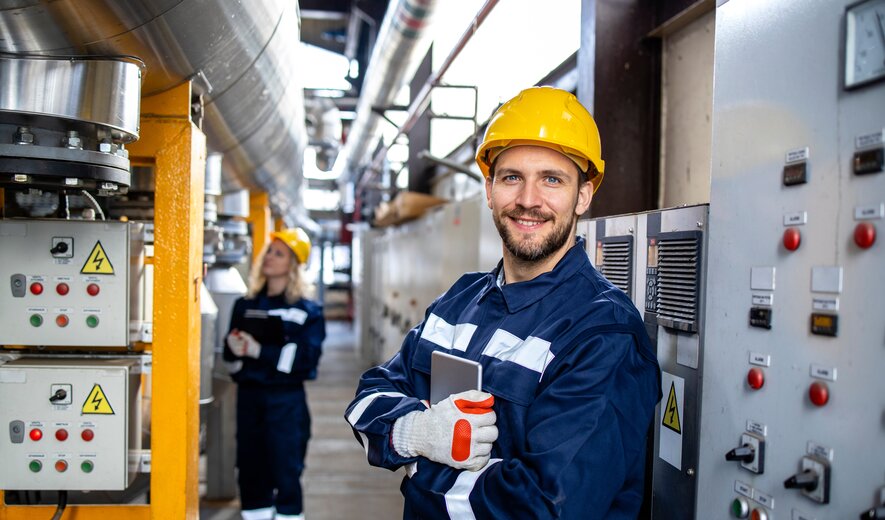 Ein Techniker in Schutzkleidung und Helm arbeitet in einer Industrieanlage und lächelt selbstbewusst in die Kamera.