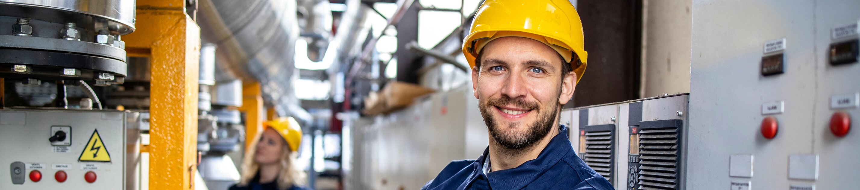 Ein Techniker in Schutzkleidung und Helm arbeitet in einer Industrieanlage und lächelt selbstbewusst in die Kamera.