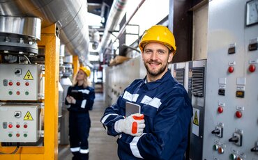 Mann mit Schutzhelm und Arbeitskleidung steht in industrieller Anlage – Symbol für technische Weiterbildung.