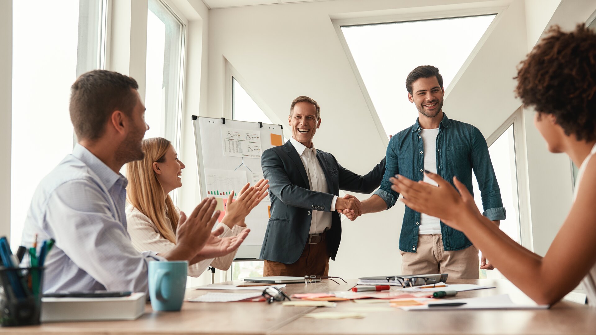 In einem hellen Besprechungsraum schütteln sich zwei Männer lachend die Hände, während das Team am Tisch applaudiert und die erfolgreiche Zusammenarbeit feiert.