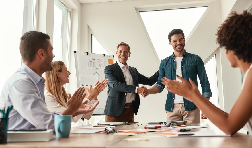 In einem hellen Besprechungsraum schütteln sich zwei Männer lachend die Hände, während das Team am Tisch applaudiert und die erfolgreiche Zusammenarbeit feiert.