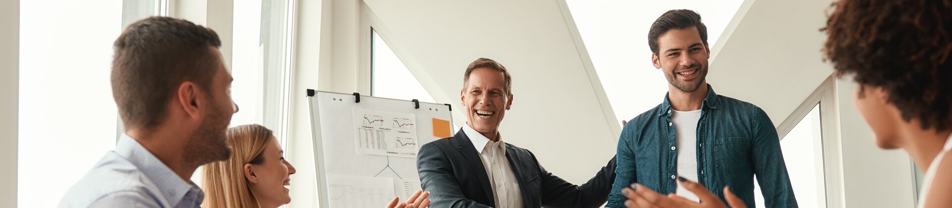 In einem hellen Besprechungsraum schütteln sich zwei Männer lachend die Hände, während das Team am Tisch applaudiert und die erfolgreiche Zusammenarbeit feiert.