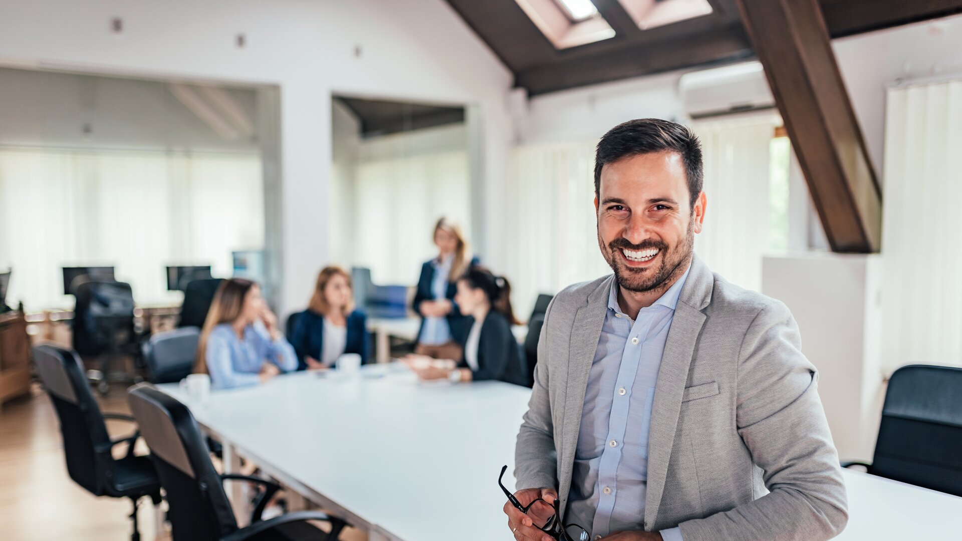 Ein freundlicher Geschäftsmann steht lächelnd im modernen Büro, während sein Team im Hintergrund in einer Besprechung zusammenarbeitet.