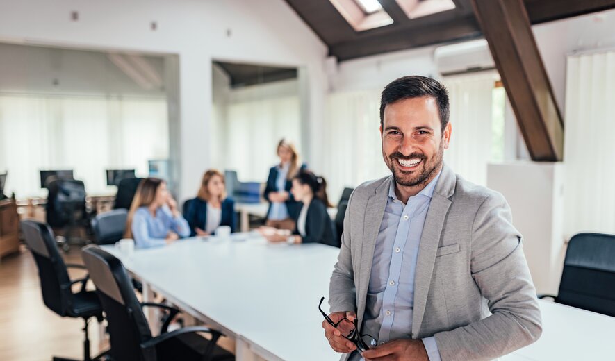 Ein freundlicher Geschäftsmann steht lächelnd im modernen Büro, während sein Team im Hintergrund in einer Besprechung zusammenarbeitet.