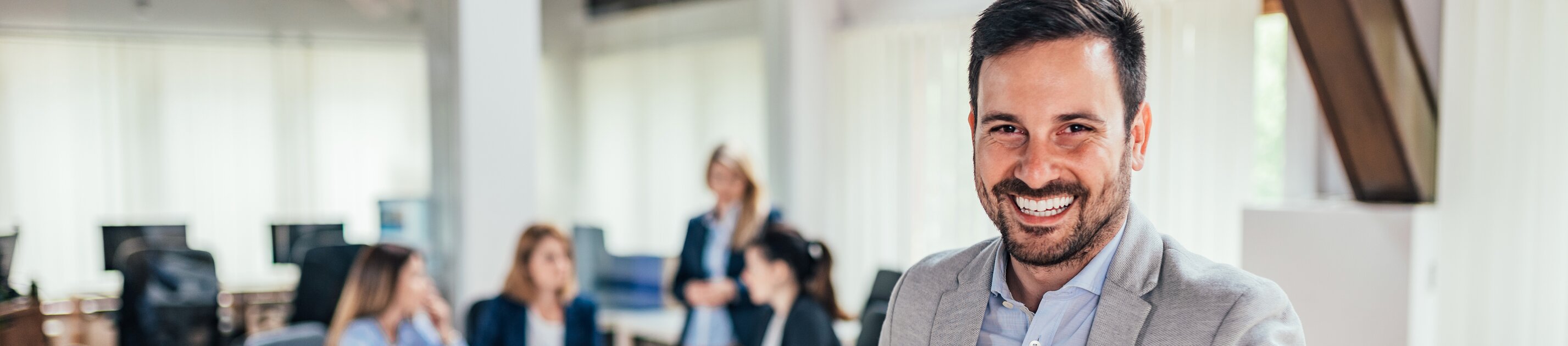 Ein freundlicher Geschäftsmann steht lächelnd im modernen Büro, während sein Team im Hintergrund in einer Besprechung zusammenarbeitet.