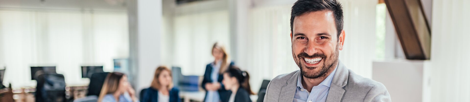 Ein freundlicher Geschäftsmann steht lächelnd im modernen Büro, während sein Team im Hintergrund in einer Besprechung zusammenarbeitet.