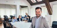 Ein freundlicher Geschäftsmann steht lächelnd im modernen Büro, während sein Team im Hintergrund in einer Besprechung zusammenarbeitet.