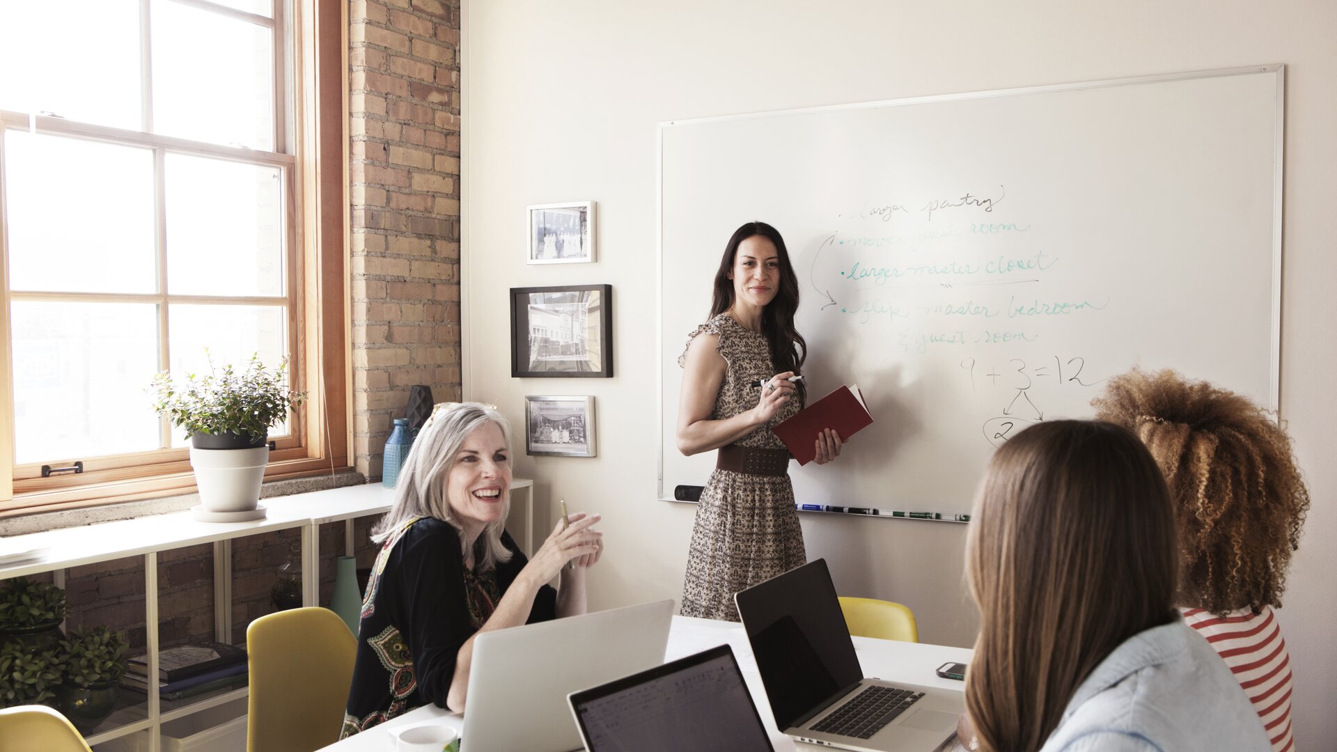 Eine Frau präsentiert Inhalte am Whiteboard, während ihr Team am Tisch aktiv mitarbeitet.