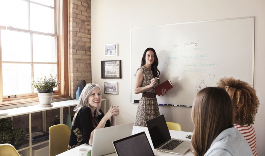 Eine Frau präsentiert Inhalte am Whiteboard, während ihr Team am Tisch aktiv mitarbeitet.