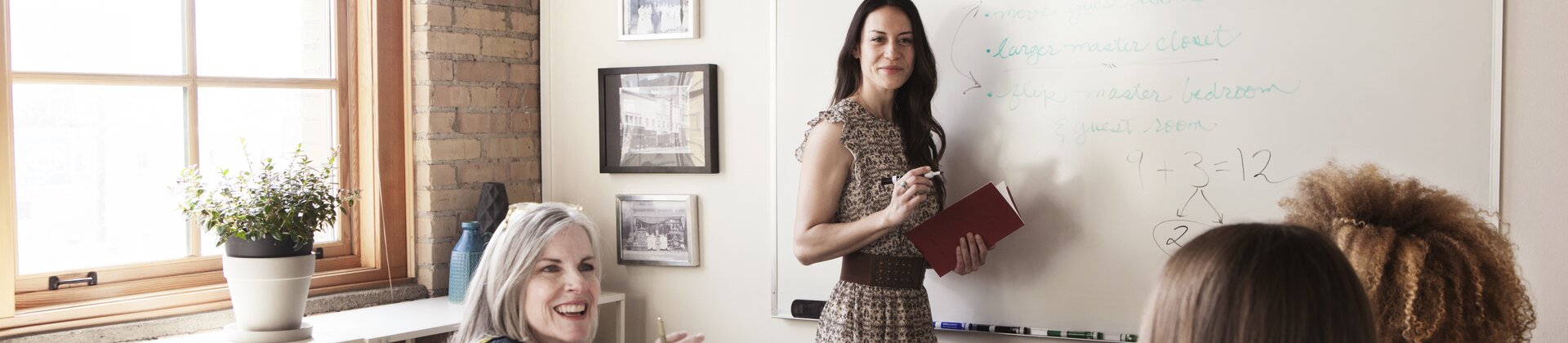 Eine Frau präsentiert Inhalte am Whiteboard, während ihr Team am Tisch aktiv mitarbeitet.