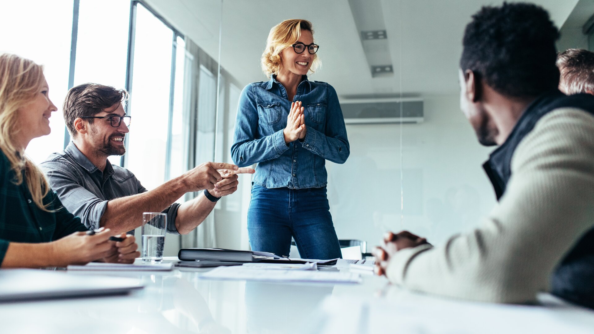 Ein lebendiges Meeting, in dem eine lachende Frau im Mittelpunkt steht und enthusiastisch zu ihrem Team spricht, während die anderen gut gelaunt reagieren.