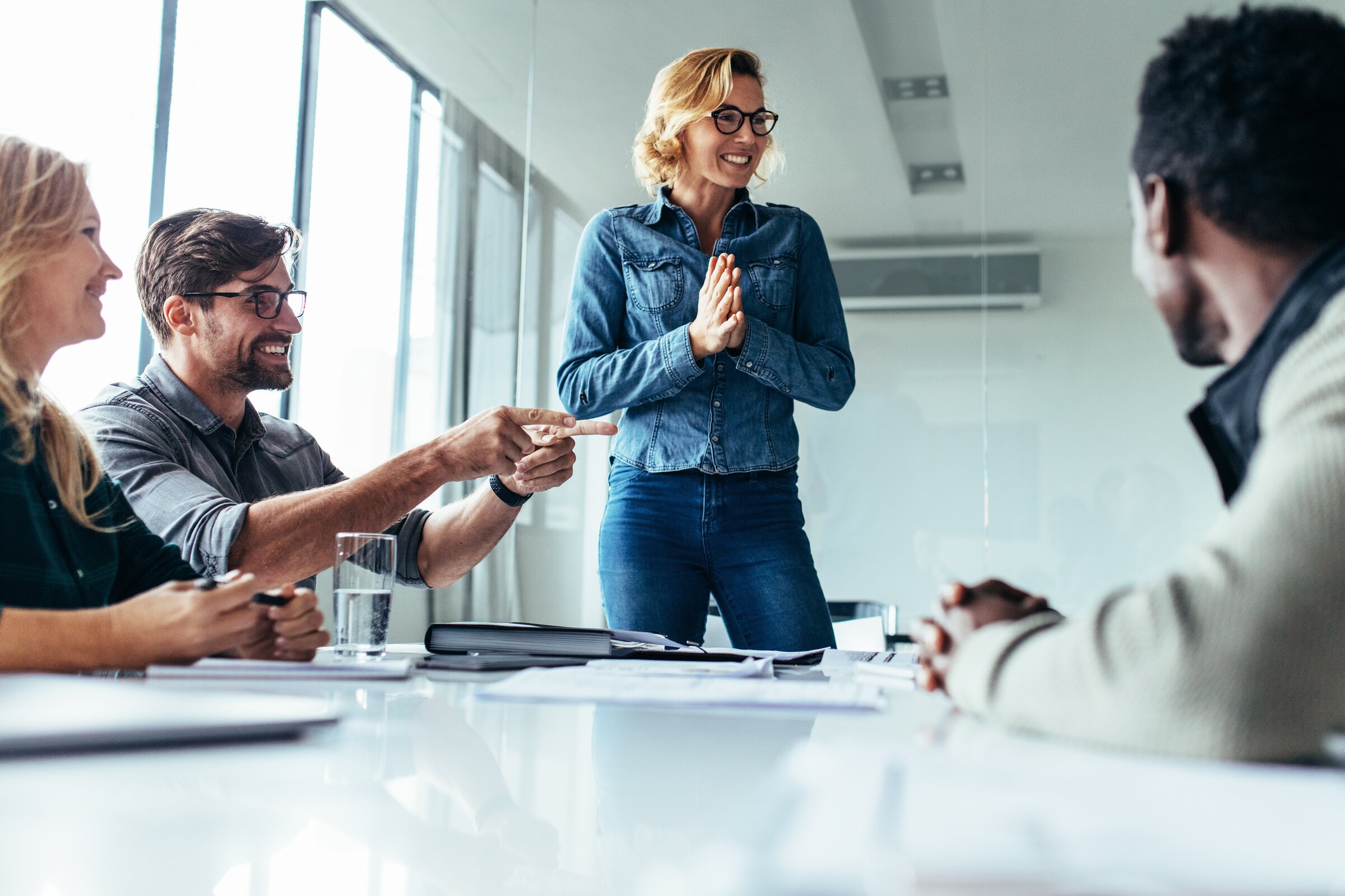 Ein lebendiges Meeting, in dem eine lachende Frau im Mittelpunkt steht und enthusiastisch zu ihrem Team spricht, während die anderen gut gelaunt reagieren.