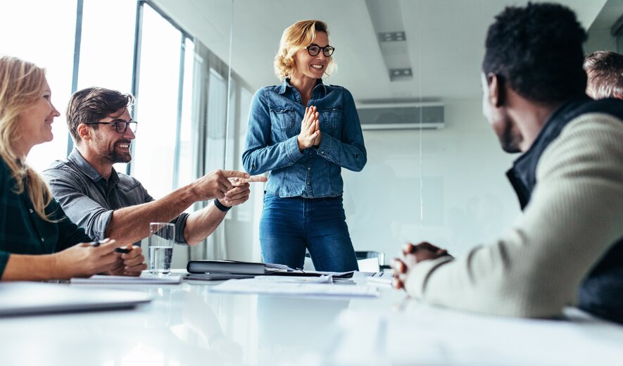 Ein lebendiges Meeting, in dem eine lachende Frau im Mittelpunkt steht und enthusiastisch zu ihrem Team spricht, während die anderen gut gelaunt reagieren.