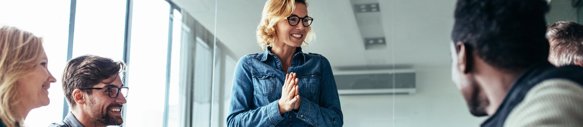Ein lebendiges Meeting, in dem eine lachende Frau im Mittelpunkt steht und enthusiastisch zu ihrem Team spricht, während die anderen gut gelaunt reagieren.