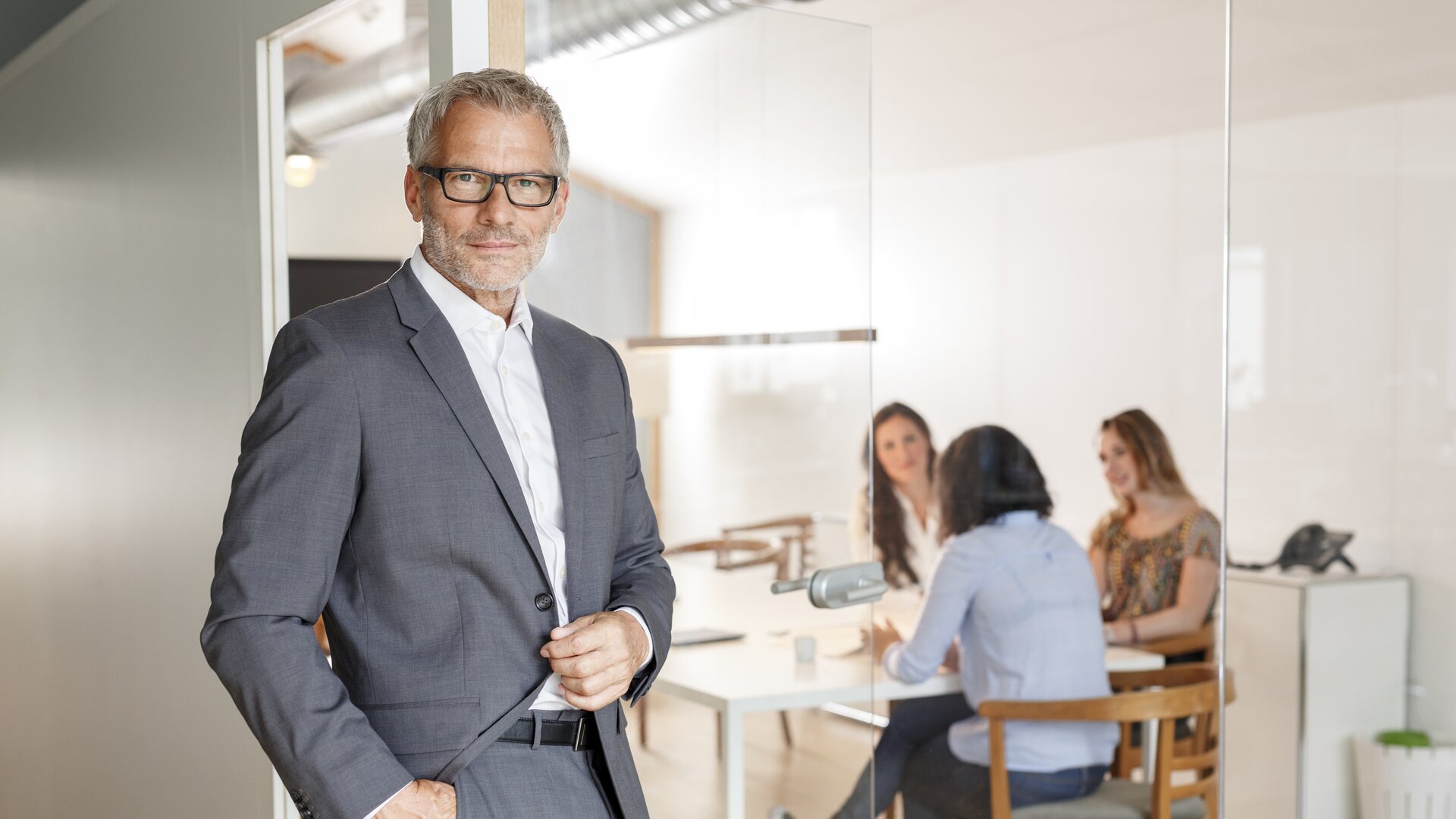 Ein elegant gekleideter Mann steht selbstbewusst vor einem Besprechungsraum, in dem mehrere Personen an einem Tisch zusammenarbeiten.