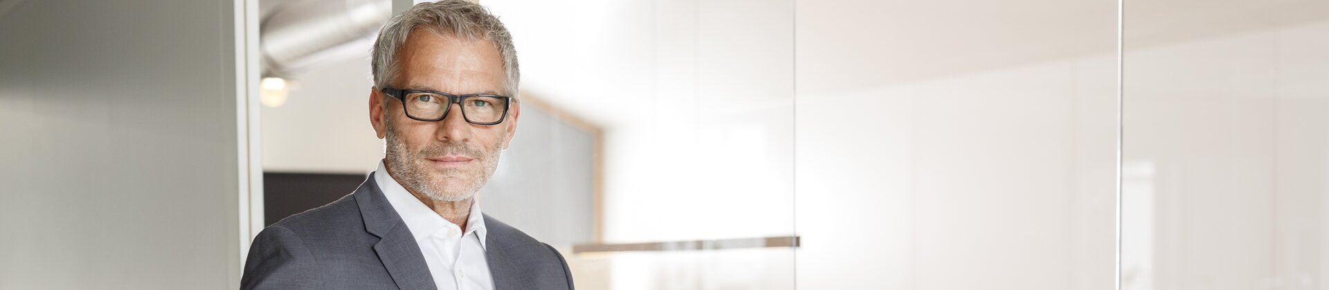 Ein elegant gekleideter Mann steht selbstbewusst vor einem Besprechungsraum, in dem mehrere Personen an einem Tisch zusammenarbeiten.