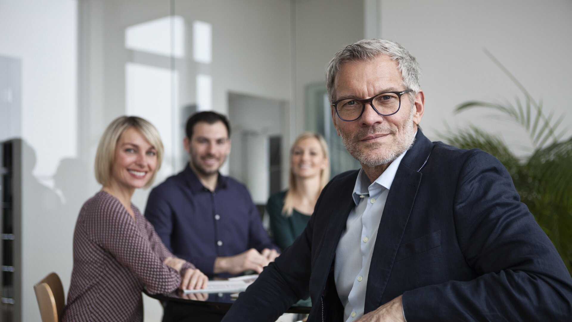 Ein souveräner Geschäftsmann sitzt im Vordergrund, während sein Team im Hintergrund freundlich in die Kamera lächelt.