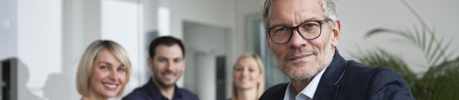 Ein souveräner Geschäftsmann sitzt im Vordergrund, während sein Team im Hintergrund freundlich in die Kamera lächelt.