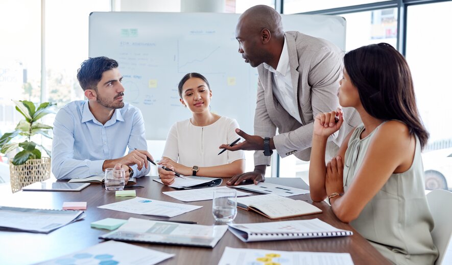 Ein Team sitzt zusammen im Meeting, während ein stehender Kollege engagiert etwas erklärt.