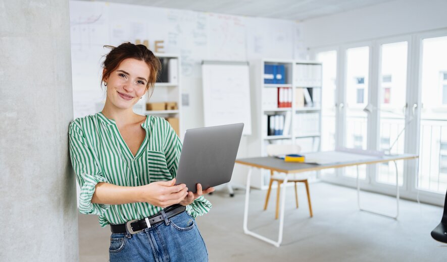Eine junge Frau lehnt entspannt an einer Säule und lächelt, während sie ein Laptop in den Händen hält.