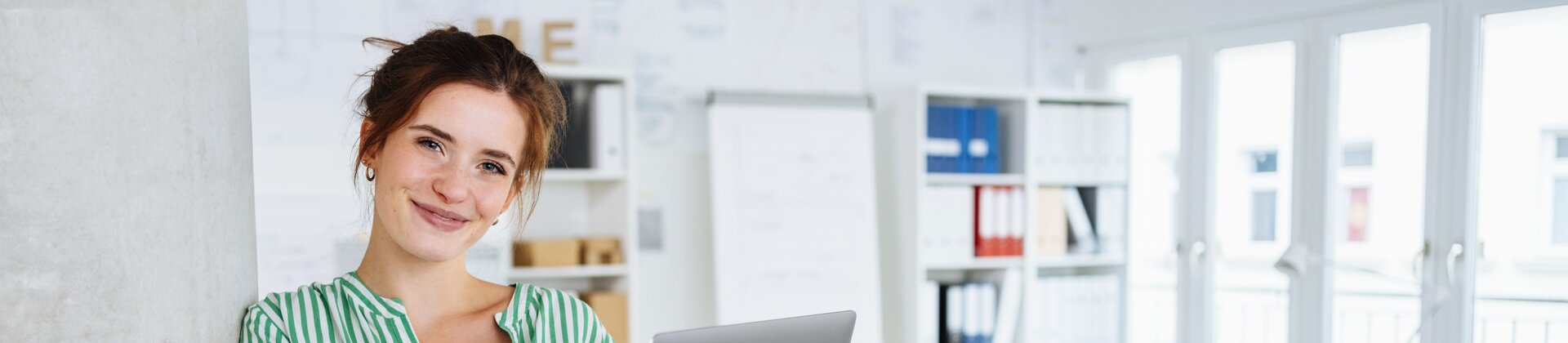 Eine junge Frau lehnt entspannt an einer Säule und lächelt, während sie ein Laptop in den Händen hält.