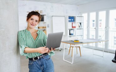 Eine junge Frau lehnt entspannt an einer Säule und lächelt, während sie ein Laptop in den Händen hält.