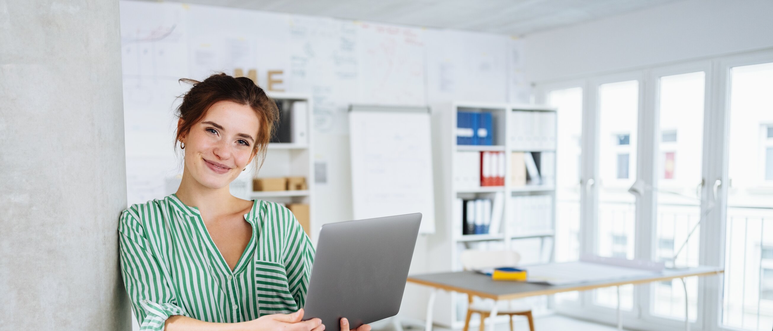 Frau lehnt lächelnd an einer Säule und hält einen Laptop in der Hand – Weiterbildung zur IHK-Fachkraft in modernem Büro.