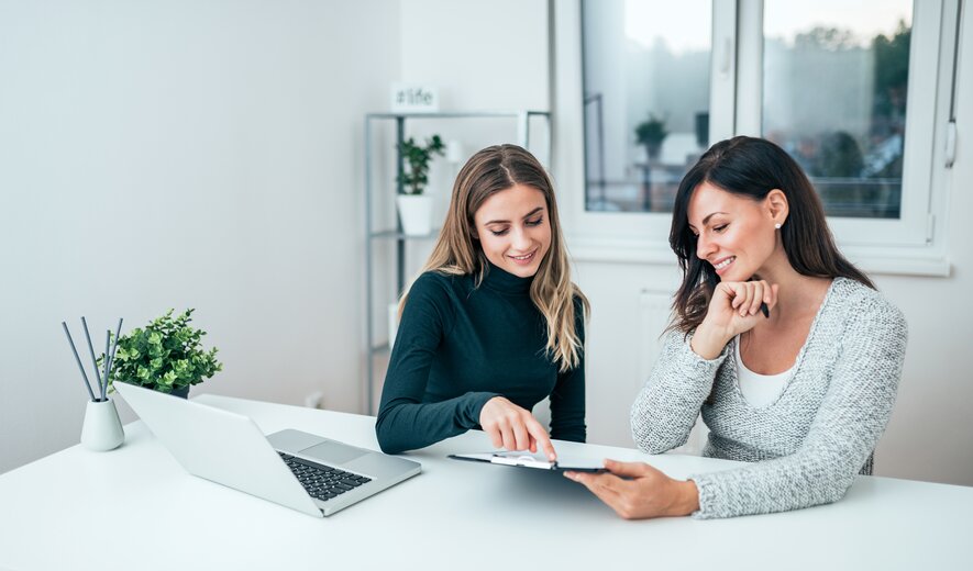 Zwei Frauen sitzen gemeinsam am Schreibtisch und besprechen lächelnd Dokumente auf einem Tablet.