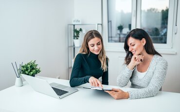 Zwei Frauen sitzen gemeinsam am Schreibtisch und besprechen lächelnd Dokumente auf einem Tablet.