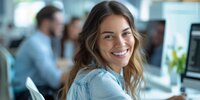Die Person auf dem Bild sitzt an einem Arbeitsplatz vor dem Computer und lächelt freundlich in die Kamera.