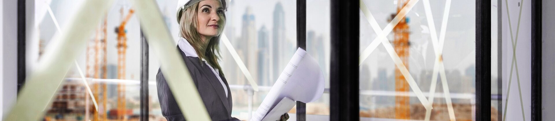 Architektin mit Bauplänen und Schutzhelm begutachtet moderne Baustelle vor Panoramafenstern.