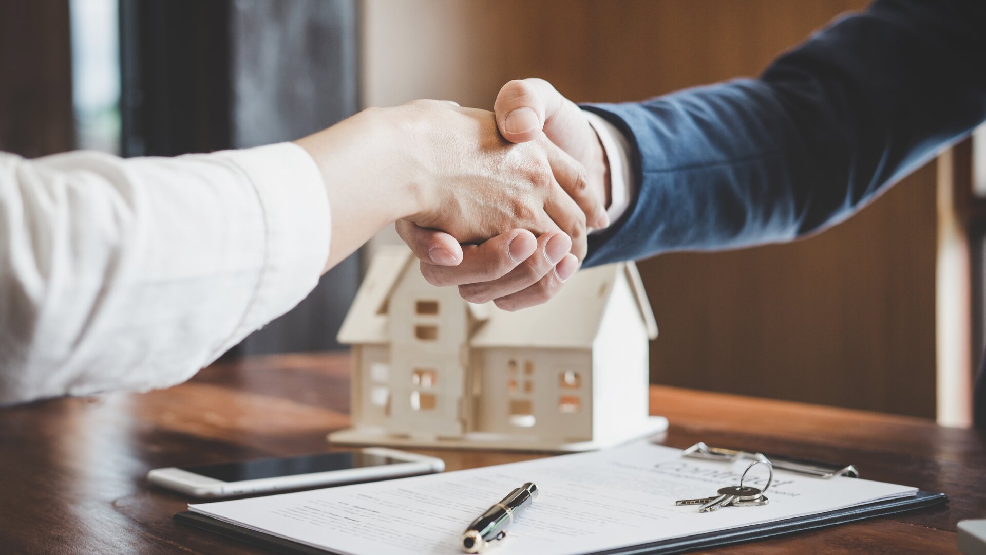 Handschlag beim Immobilienkauf mit Vertrag, Schlüssel und Hausmodell auf dem Tisch