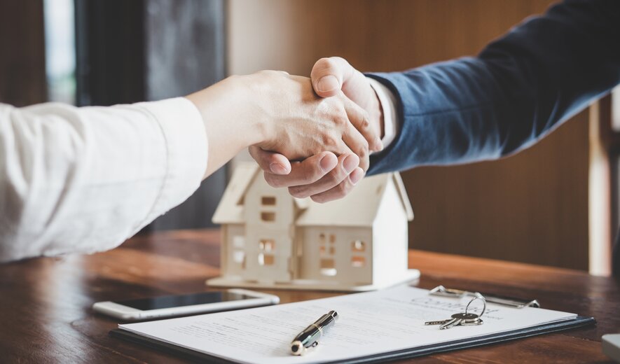 Handschlag beim Immobilienkauf mit Vertrag, Schlüssel und Hausmodell auf dem Tisch