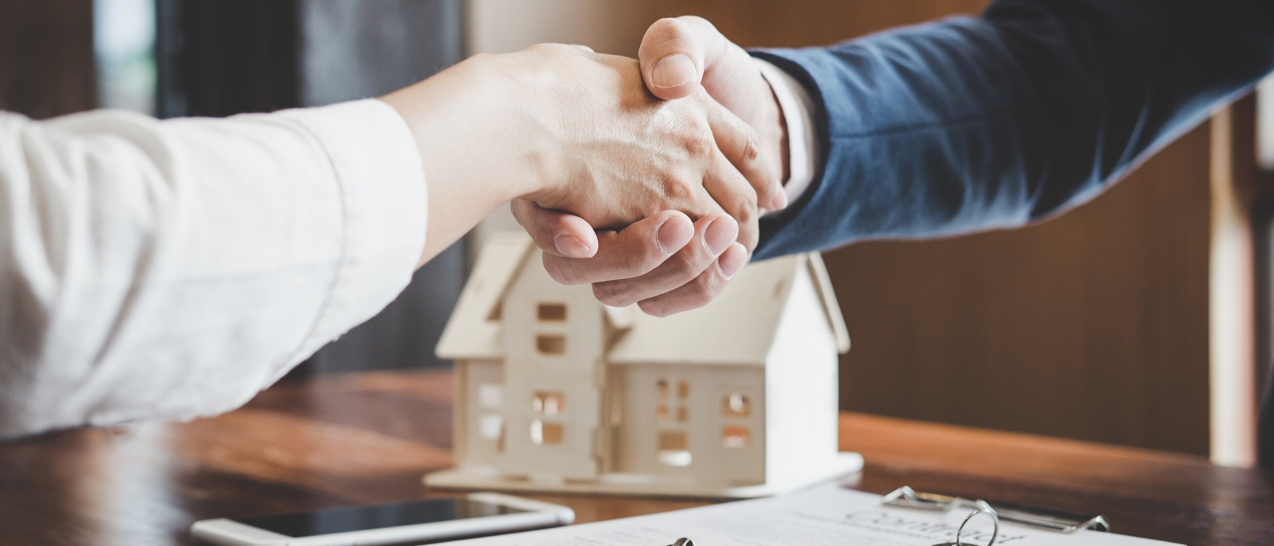 Weiterbildung im Bereich Immobilienverwaltung: Zwei Personen reichen sich die Hand über einem Vertragstisch, auf dem ein Modellhaus und ein Schlüsselbund liegen.