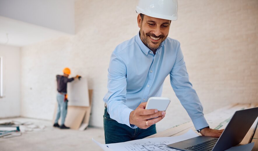 Der Mann auf dem Bild lächelt, während er auf einer Baustelle mit Smartphone und Laptop arbeitet.