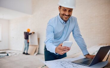 Bauleiter mit Schutzhelm prüft Baupläne und Smartphone auf der Baustelle – Weiterbildung für Bauträger