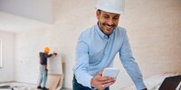 Der Mann auf dem Bild lächelt, während er auf einer Baustelle mit Smartphone und Laptop arbeitet.