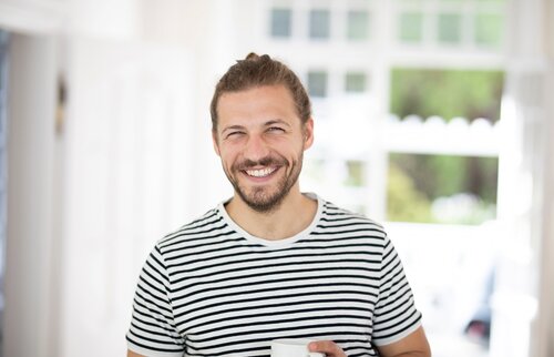 Lächelnder Mann in gestreiftem T-Shirt hält eine Tasse in heller, freundlicher Wohnumgebung