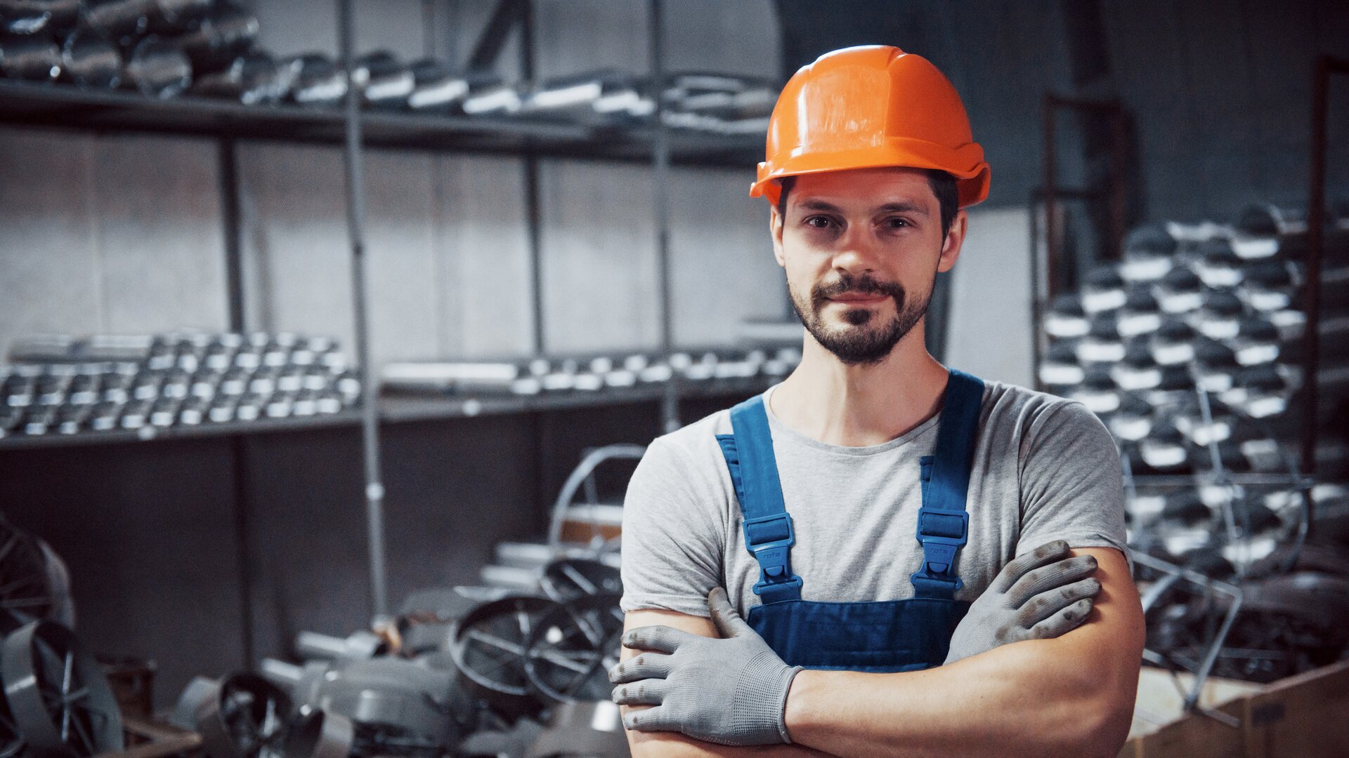 Industriearbeiter mit Schutzhelm und Handschuhen steht in Werkhalle mit Metallteilen