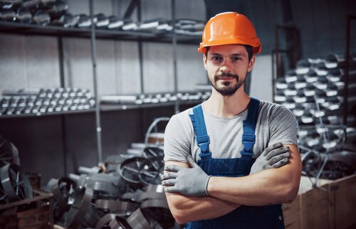 Industriearbeiter mit Schutzhelm und Handschuhen steht in Werkhalle mit Metallteilen