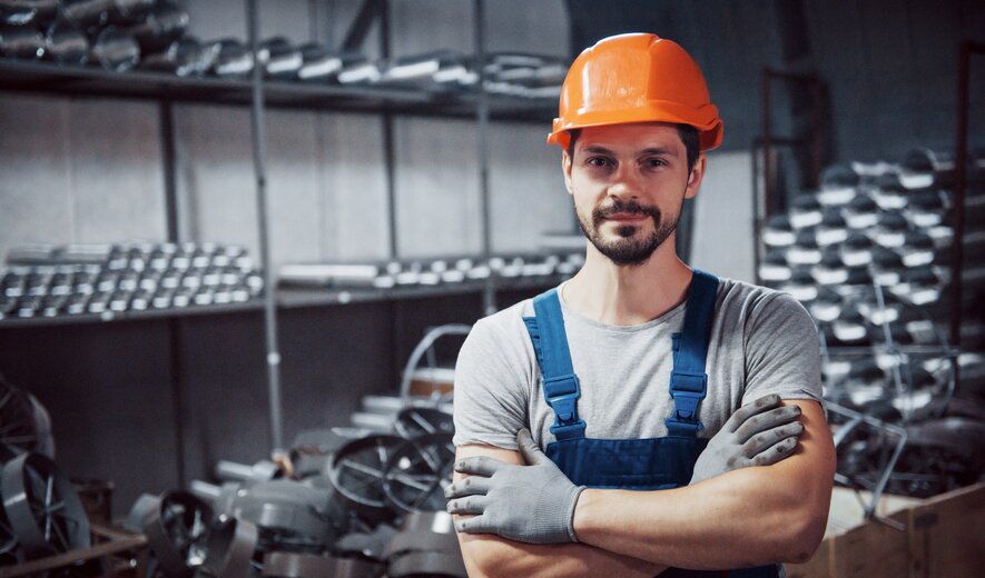 Industriearbeiter mit Schutzhelm und Handschuhen steht in Werkhalle mit Metallteilen